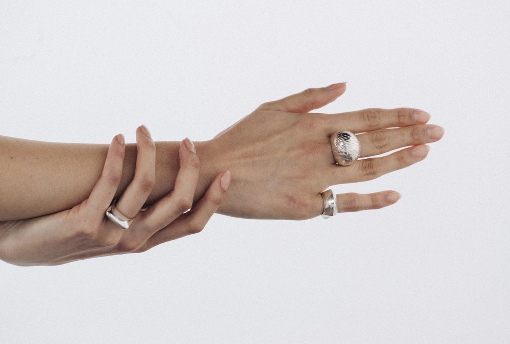 Two hands shown against a plain light background, each wearing a silver-toned ring. One ring has a large clear oval stone, and the other is a simple band ring.