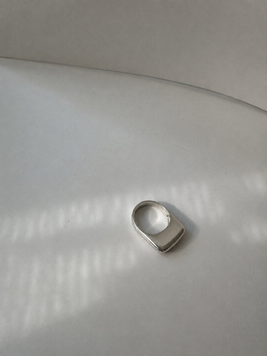 A silver-toned metal ring with a wide, rounded rectangular face, photographed on a light gray surface with soft window-box style shadows.