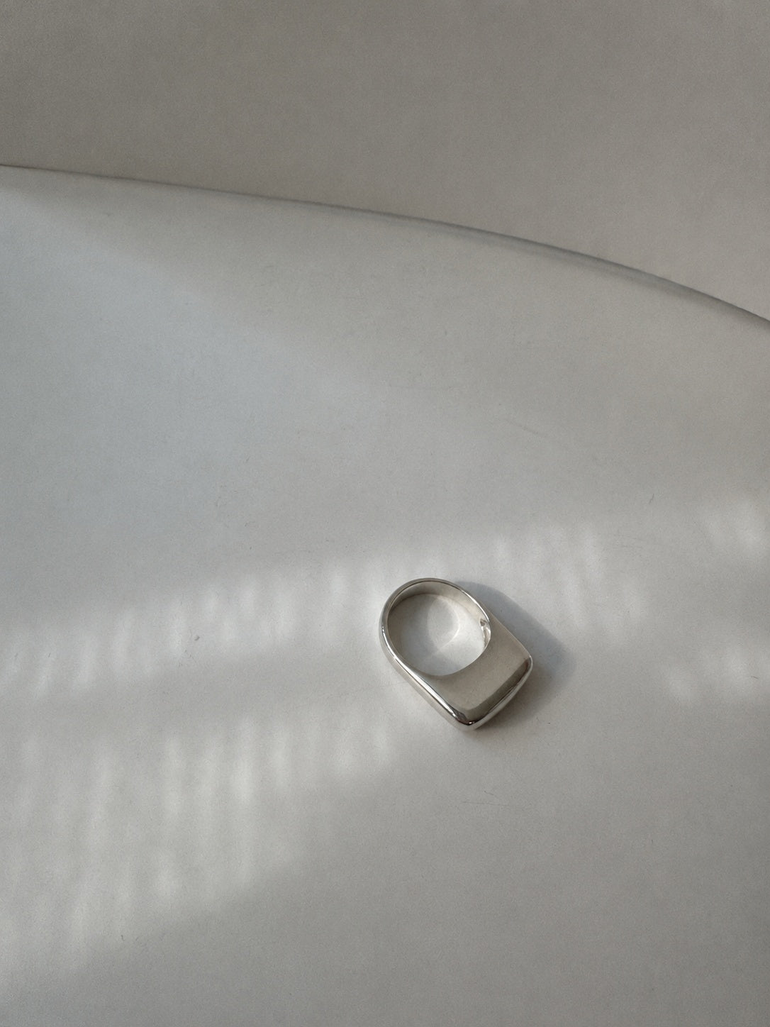 A silver-toned metal ring with a wide, rounded rectangular face, photographed on a light gray surface with soft window-box style shadows.