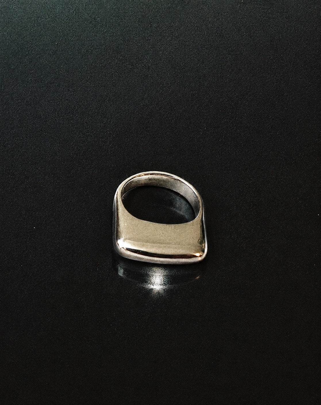 A polished silver signet ring with a wide, rounded rectangular face, displayed on a dark reflective surface.