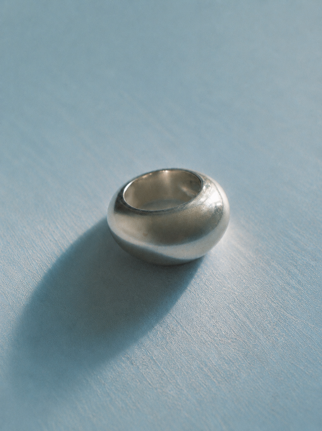 A polished silver ring with a smooth, rounded, dome-like shape resting on a light blue textured surface with a soft shadow.